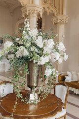 Floral arrangement of white roses, ivy, and green herbs in old high metal cup on round table . 