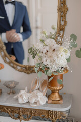 Elegant wedding shoes and snow-white bridal bouquet in vintage vase stand on old wall table.