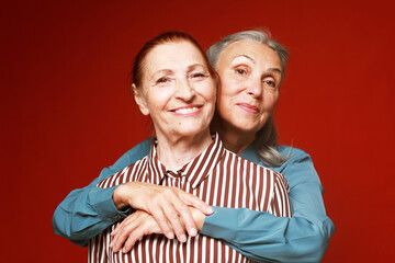 Two elderly women friends hugging on red background.