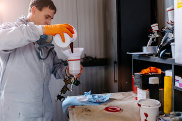 Male car painter in protective clothes pouring mixed paint from a can into a plastic mixing bucket...