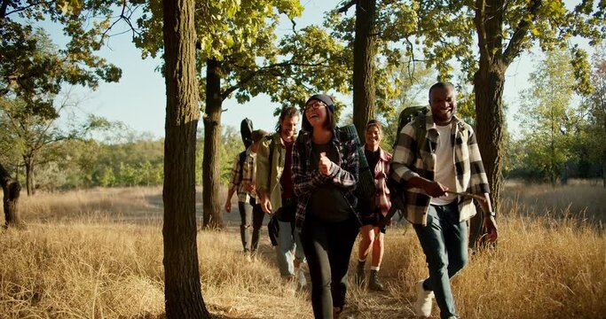 Front view of a happy group of several pairs of people in special hiking clothes with hiking backpacks walking through the forest near dried yellow summer grass. A Black man talks to a blonde girl