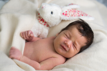 Fototapeta na wymiar Portrait of a cute newborn boy under a white blanket, a white toy bunny lies next to him. Close-up