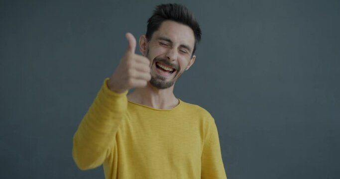 Slow motion portrait of happy young man showing thumbs-up hand gesture expressing admiration and approval on gray background. Emotion and people concept.