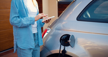 Close up of woman hands holding smartphone. Girl plugs power cable to charge electric car in parking near home. Eco friendly alternative energy green environment concept.