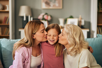 In a heartwarming snapshot, a young girl finds herself enveloped in love as her mother and grandmother affectionately kiss her cheeks. Her eyes are closed in delight, her mouth open in a joyful giggle