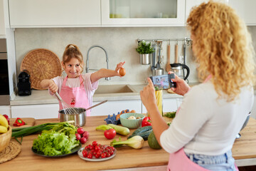A spirited young girl excitedly displays an egg while whisking a mixture, as her mother captures the spontaneous act with her camera