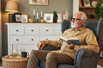 Elderly man sitting in armchair and reading a book at home - Powered by Adobe