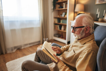 Elderly man sitting in armchair and reading a book at home