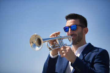 Young Hispanic man, wearing a jacket and sunglasses, playing a pretty, silvery trumpet outdoors....