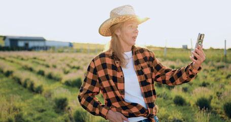 Middle-aged beautiful woman farmer in a hat has online conference with agribusiness partners standing in a field at sunset. Agricultural specialist video call by phone.