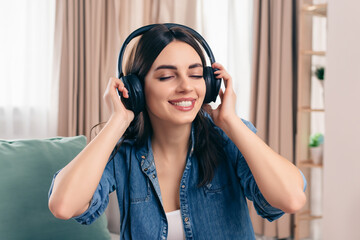 beautiful girl sits on the sofa near the window and listens to music in large headphones, smiling girl has fun with music