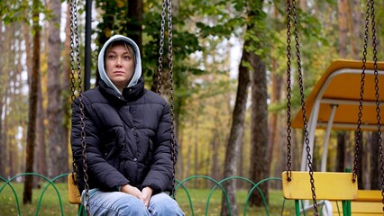 A beautiful young thoughtful woman is sad on a swing in a city park. A woman in warm clothes and a hood looks thoughtfully into the distance, worried about problems, depression and loneliness.