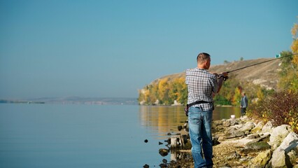 Fototapeta na wymiar A mature male fisherman in casual clothes casts a lure with a spinning rod while fishing on the river bank. Profile view of an adult man spinning fishing in the weekends. Copy space.