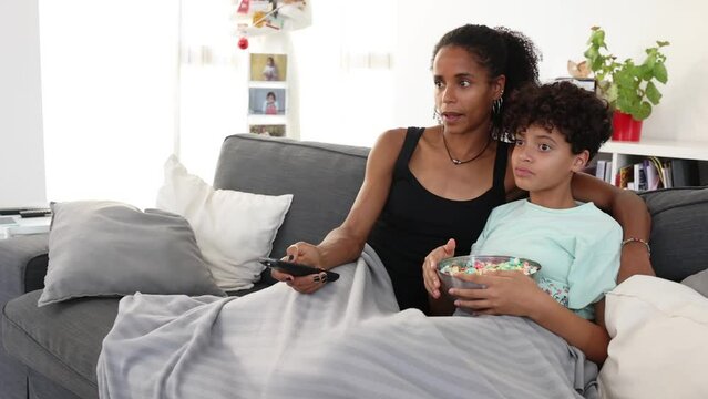 Smiling mother and daughter watching tv and eating popcorn on a couch of a living room. Mother and daugther sharing a good moments at home while wathing a movie.