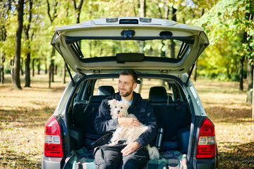 Young man with a dog relaxing and enjoying nature sitting in car trunk