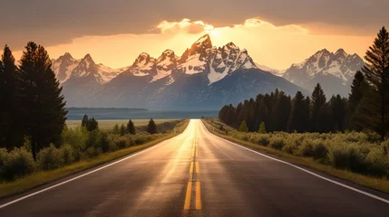 Fotobehang Tetongebergte An open road leads to the Grand Teton's mountain range, rising in the distance beyond a thick pine forest. The last rays of sunlight shine on the mountain. Photo shot vertically to include more road.