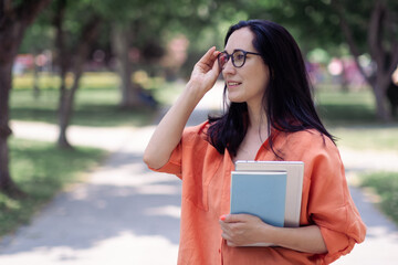 Business lady in stylish casual clothes adjusting glasses standing in trees shade in green park teacher holding paper notebook and tablet in spring urban garden