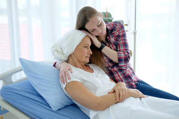 Caucasian daughter is Holding hands Elderly Mother in white headscarf is laying on bed in hospital...