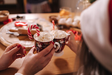 Winter drink, white mug with marshmallows in females hands in white sweater. Home bakery, cooking traditional festive sweets. Gingerbread dough on wooden table. New Year celebration traditions.