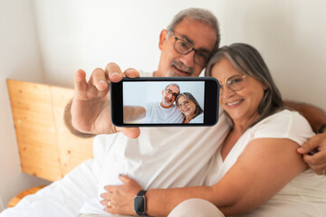 Happy old european husband and wife make selfie on phone screen on bed in bedroom