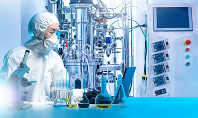Scientist microbiologist. Laboratory assistant with microscope and laptop. Bioreactor near female scientist. Biologist in protective suit and mask. Scientist sitting at table with test tubes
