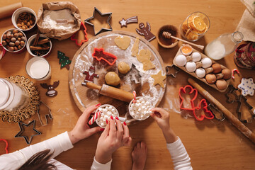 Top view of winter drink, white mug with marshmallows in females hands in white sweater. Home bakery, cooking traditional festive sweets. Gingerbread dough on wooden table. New Year traditions.