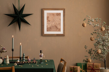 Aesthetic composition of christmas dining room interior with mock up poster frame, table, green tablecloth, candle with candlestick, brown wall, gifts, and personal accessories. Home decor. Template.	