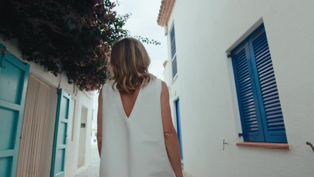 Cinematic following shot of young woman explore small quaint town on mediterranean coast. Pretty and beautiful female tourist look at camera on white spanish village street