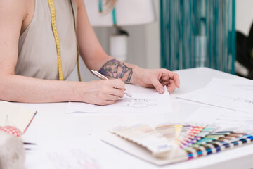 Lady dressmaker with tattoo draws clothes model on paper at desk in fashion atelier tailor creates pencil sketch of stylish outfit in sewing studio closeup