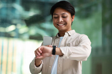 Smiling young asian man checks time on smartwatch outdoor