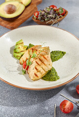 Close-up vertical side view of grilled chicken fillet with avocado and spinach on a ceramic plate. The grey concrete background with hard shadows emphasizes the dish's healthy appeal