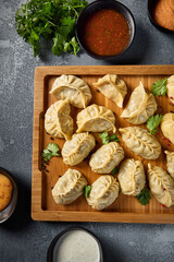 Vertical top view image of manti and eastern dumplings assortments, served on a wooden board. The accompanying sauces, vibrant and rich, become a focal point, beautifully contrasting the gray