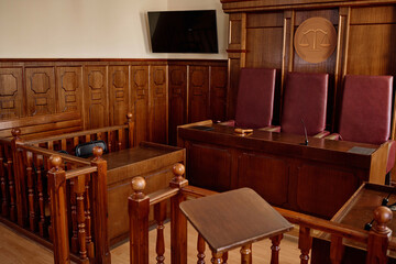 Corner of spacious courtroom with screen, wooden desks and railings and leather chairs standing in row along wall with symbol of justice