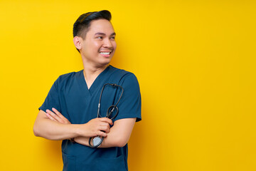 Professional young Asian man doctor or nurse wearing a blue uniform standing confidently while holding stethoscope and crossed hands, smiling friendly aside isolated on yellow background