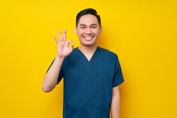 Professional young Asian male doctor or nurse wearing a blue uniform showing an okay sign, looking at camera with smile isolated on yellow background. Healthcare medicine concept