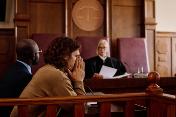 Young anxious male suspect covering his face by hands while sitting in front of camera against judge passing judgement during trial session