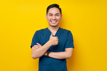 Professional young Asian male doctor or nurse wearing a blue uniform showing thumb up or like, looking at camera with smile isolated on yellow background. Healthcare medicine concept - obrazy, fototapety, plakaty