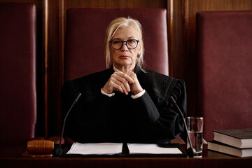 Mature impartial female judge in eyeglasses and black mantle looking at camera while sitting in front of participants of trial session in courtroom