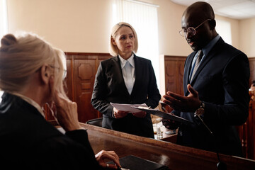 Defense and prosecution sides of court case standing in front of mature female judge while African...