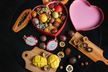 An assortment of fresh exotic fruits laid out on a black table, wooden planks. Some fruits are cut and peeled.