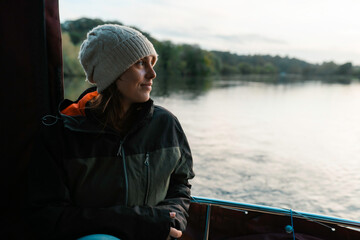 Woman on a river boat cruise, smiling and sailing, wearing a coat and wool hat, Norfolk Broads,...