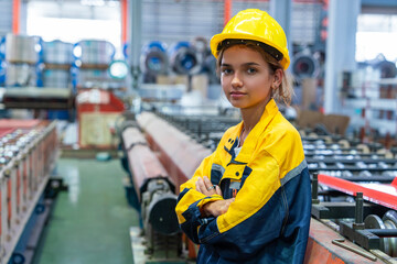 Latin female engineer wearing a uniform shirt and  helmet stands with her arms crossed over her chest, operating a machine, watching a steel roof production line in a factory.