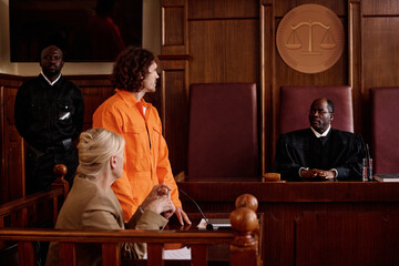 Side view of suspect in orange jumpsuit and his attorney looking at mature African American impartial judge while prisoner speaking to him