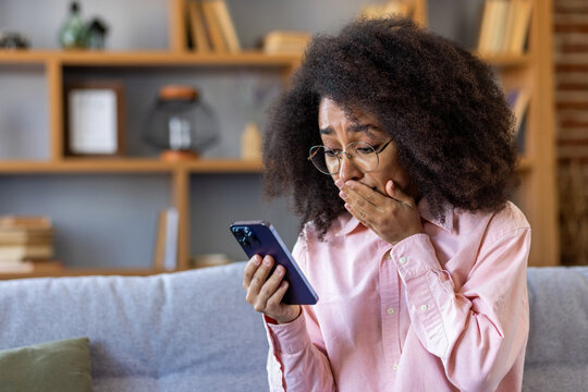Sad disappointed woman received breaking news on phone, african american woman reading bad news online using app on smartphone, sitting at home on sofa in living room of house.