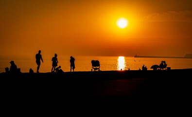 A line of families and holidaymakers on the beach at Seaford, East Sussex, silhouetted in black against a red sky and setting sun.