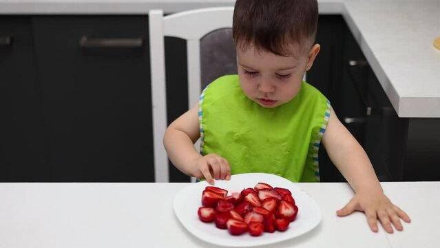 slice of strawberries fruit on plate and baby boy kid is refusing to eat. red strawberry, vitamins healthy nutrition. child with bib in kitchen 4k