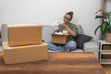 Blank mock-up parcel boxes on a table, with a woman on the backdrop engaged in unboxing her online shopping delivery, encapsulating the essence of shipping and marketplace shopping.
