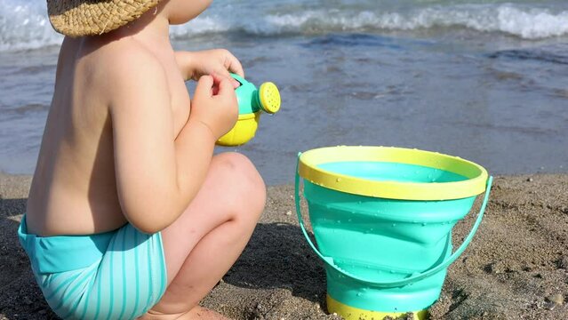 adorable baby boy playing on beach sea side with toy watering can and bucket.kid with sun cap on head sitting on sands or with feet in water. waves hitting legs.summer time vacation concept 4k