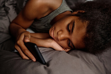 Youthful African American girl keeping head on palm while lying in bed late at night and reading message containing harassment stuff