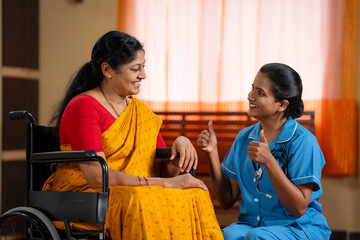 Joyful Indian nurse building confidence or encouraging recovered sick woman on wheelchair at home -...